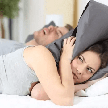 a woman covering her head with a pillow while the man sleeping next to her is snoring