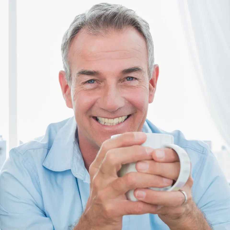 Middle-aged man with gray hair smiles at the camera while holding a white mug with both hands. He is wearing a light blue shirt and sitting indoors.