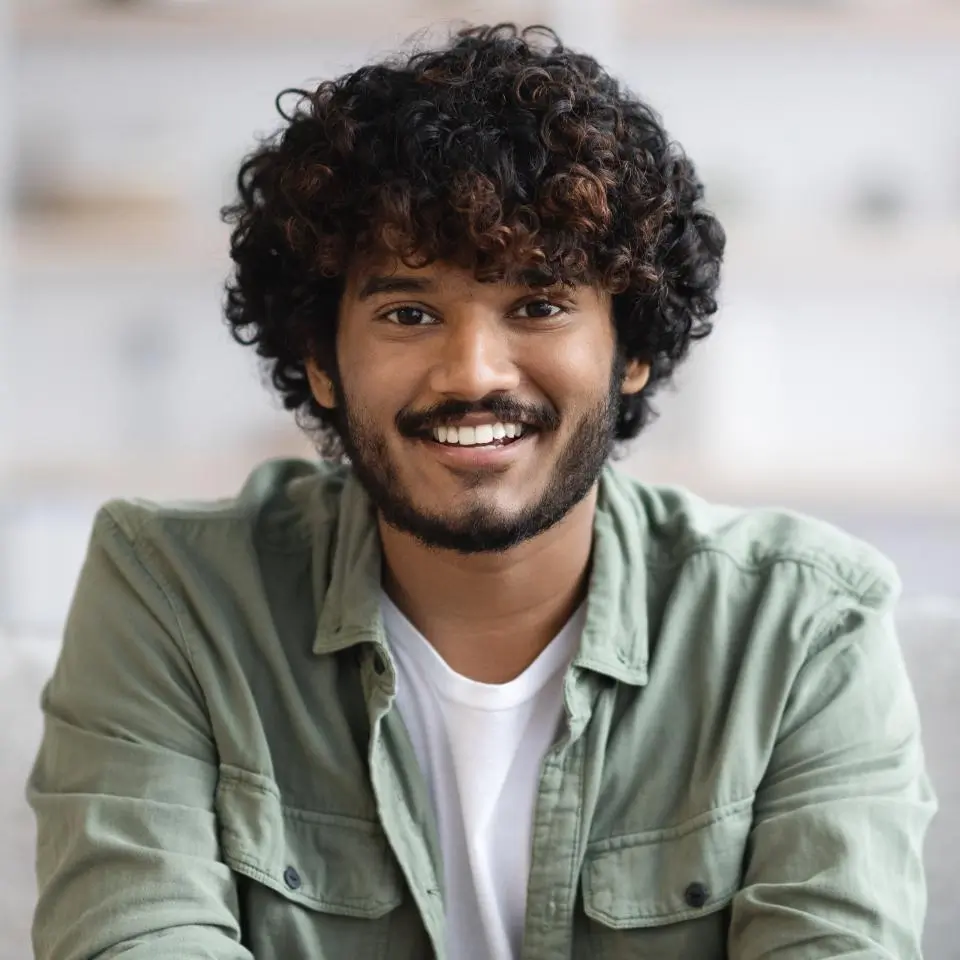 A young man with curly hair and a beard, wearing a green button-up shirt over a white t-shirt, smiling while sitting indoors.