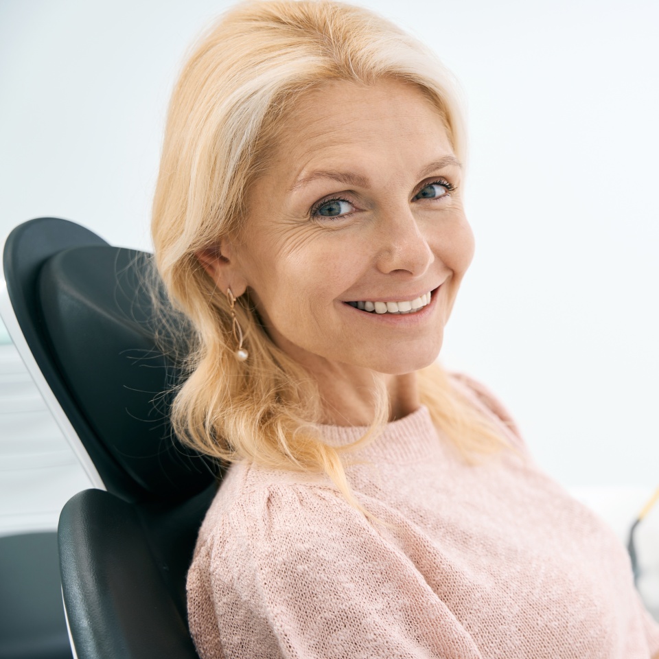 A woman with blonde hair sits in a dental chair, smiling at the camera. She is wearing a light pink sweater in a bright, clean office setting.