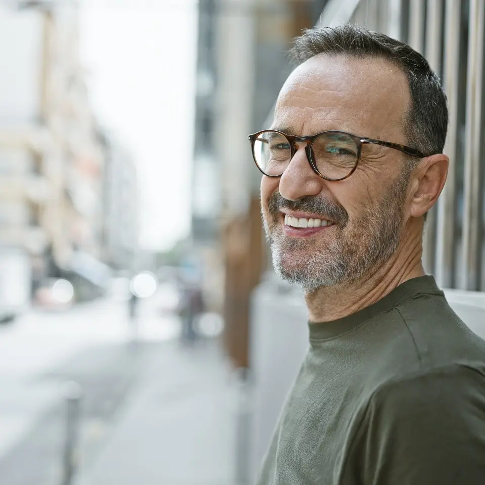A middle-aged man with glasses and a beard smiles while standing on a city sidewalk, wearing a green shirt.